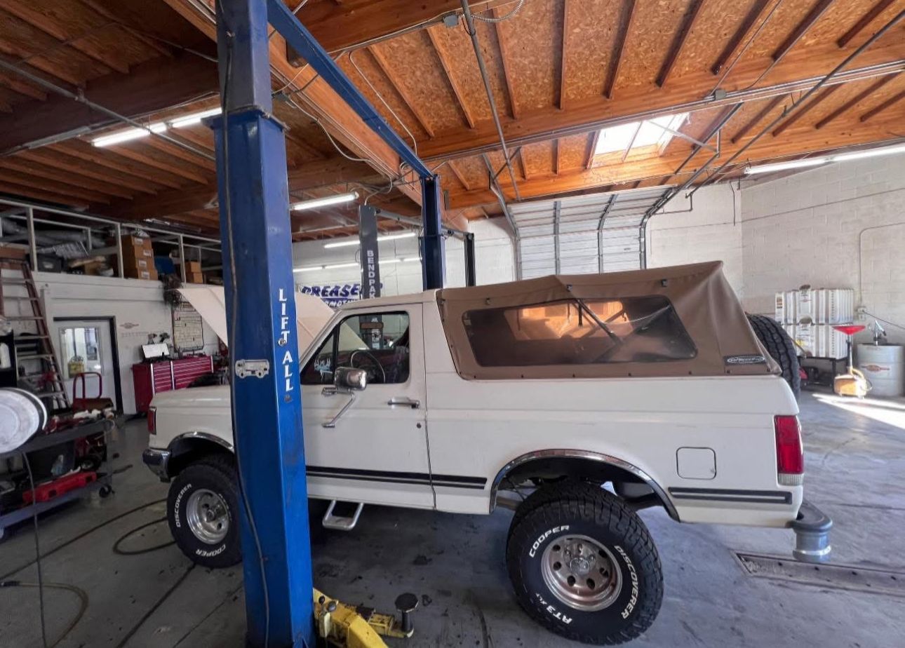 White Ford Bronco in a garage, lifted on a car lift. The soft top is tan | Greasers Automotive