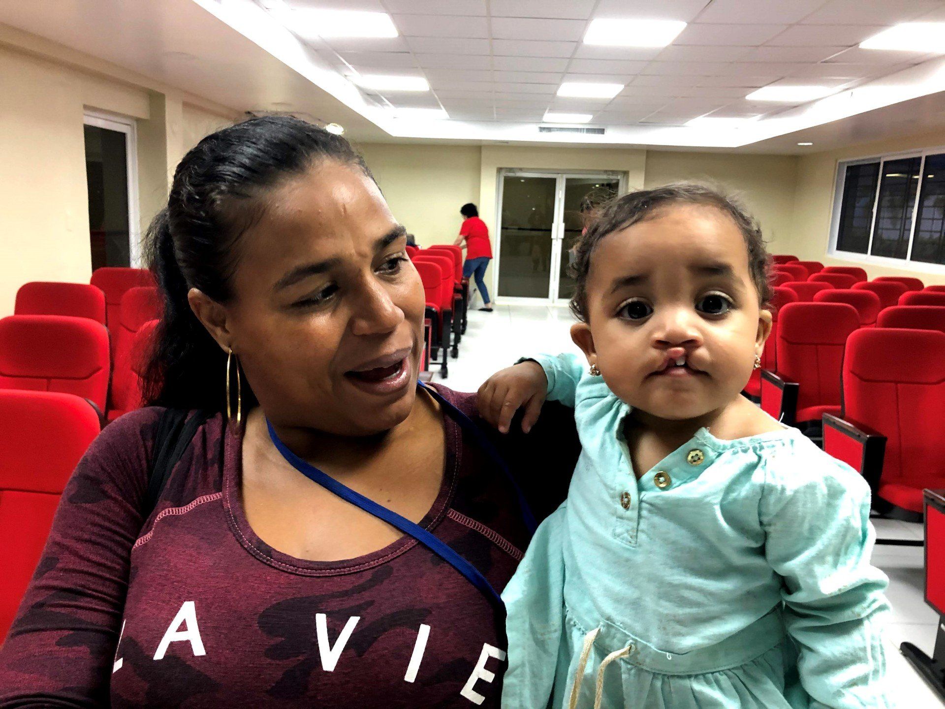 A woman is holding a baby in a room with red chairs.