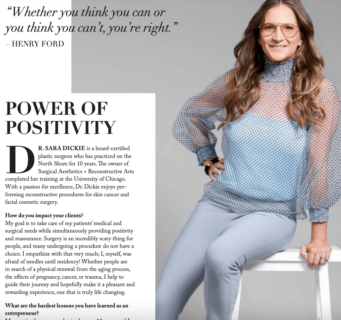 A woman wearing glasses is sitting on a stool in front of a sign that says power of positivity