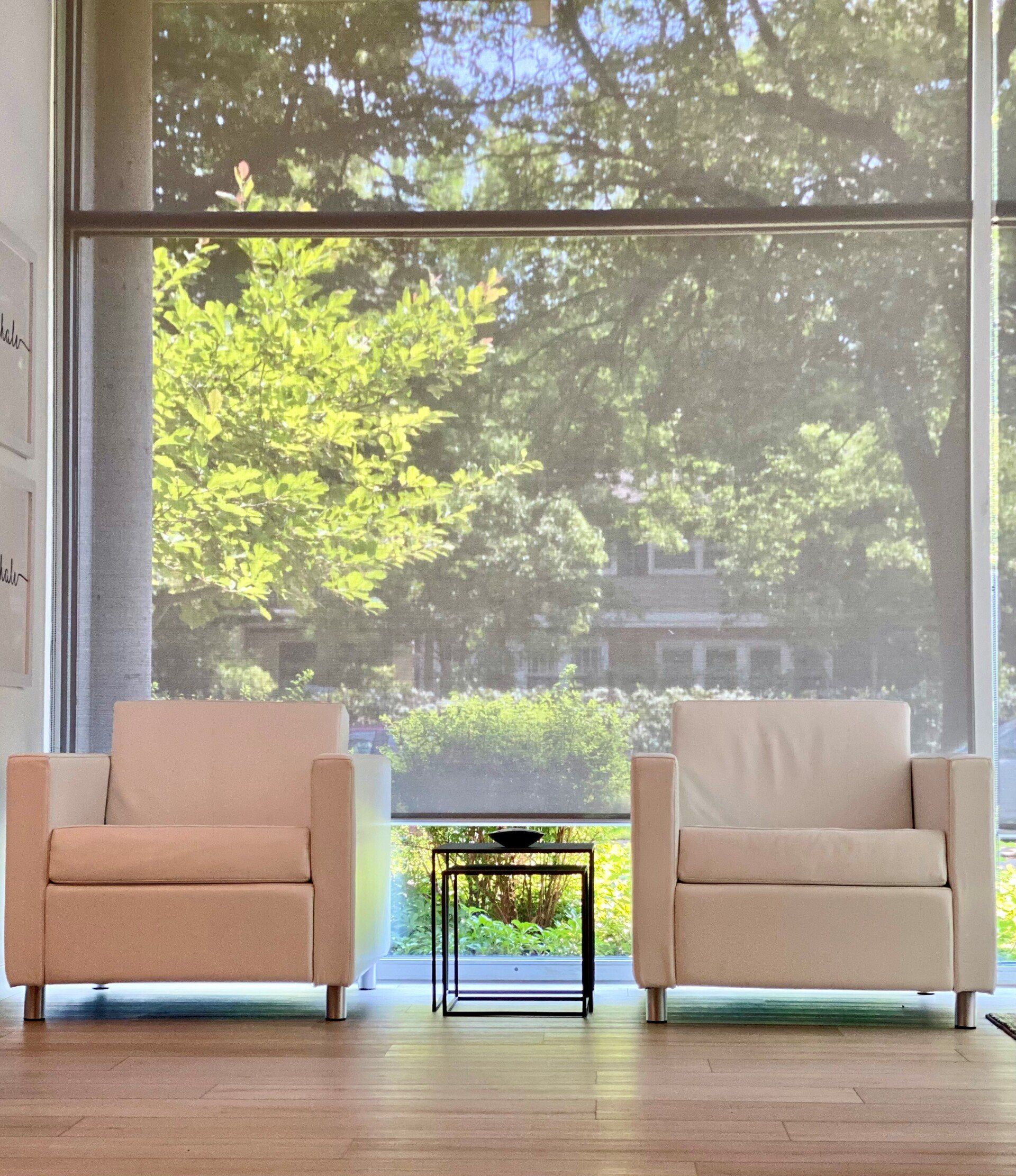Two white chairs in front of a large window with trees in the background