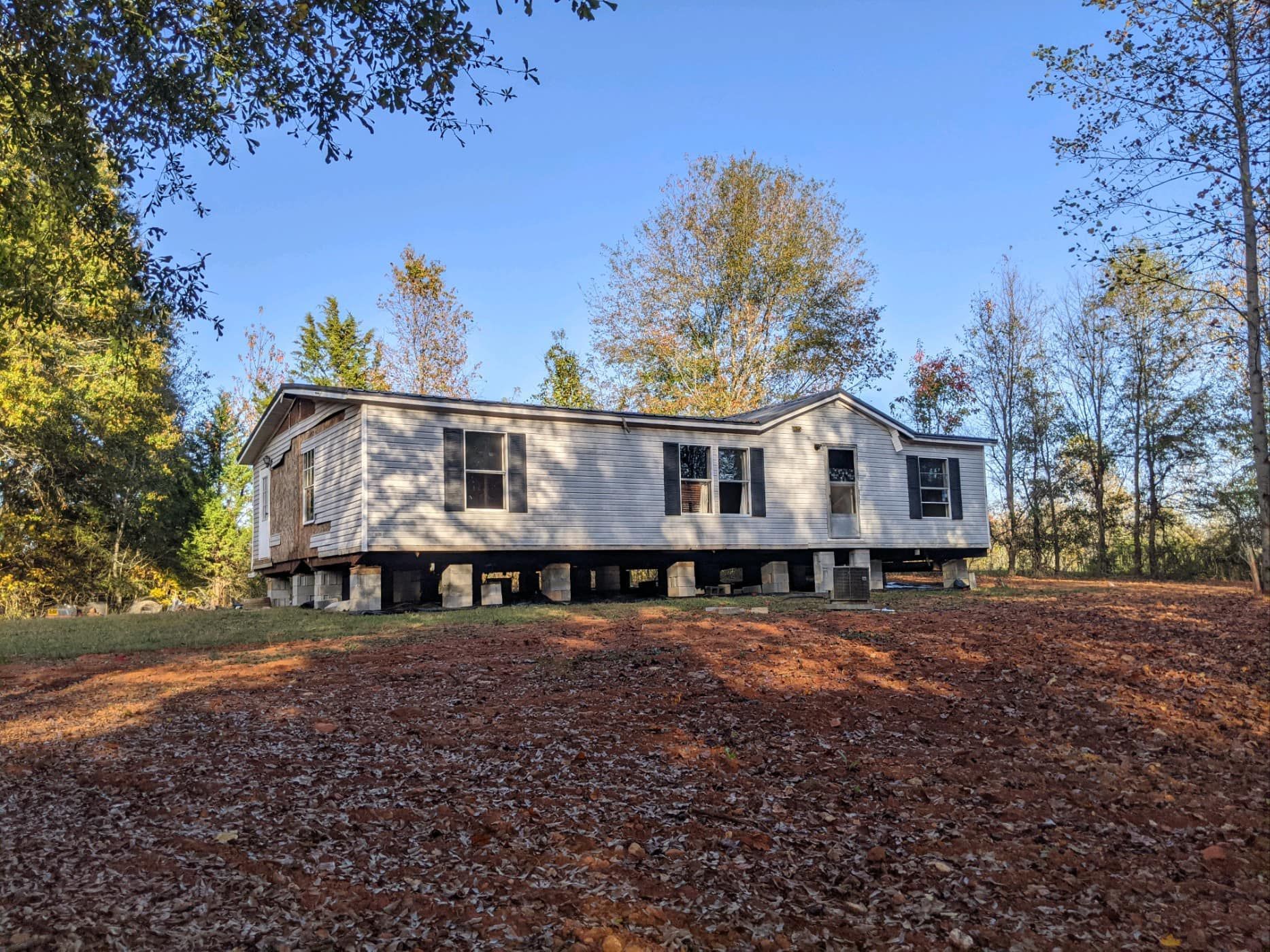 A mobile home is sitting on top of a dirt field surrounded by trees.