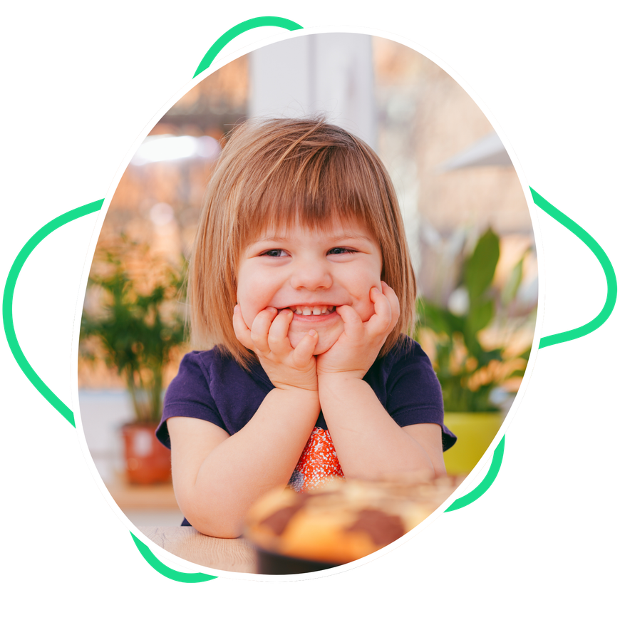 A little girl is sitting at a table with her hands on her chin and smiling.