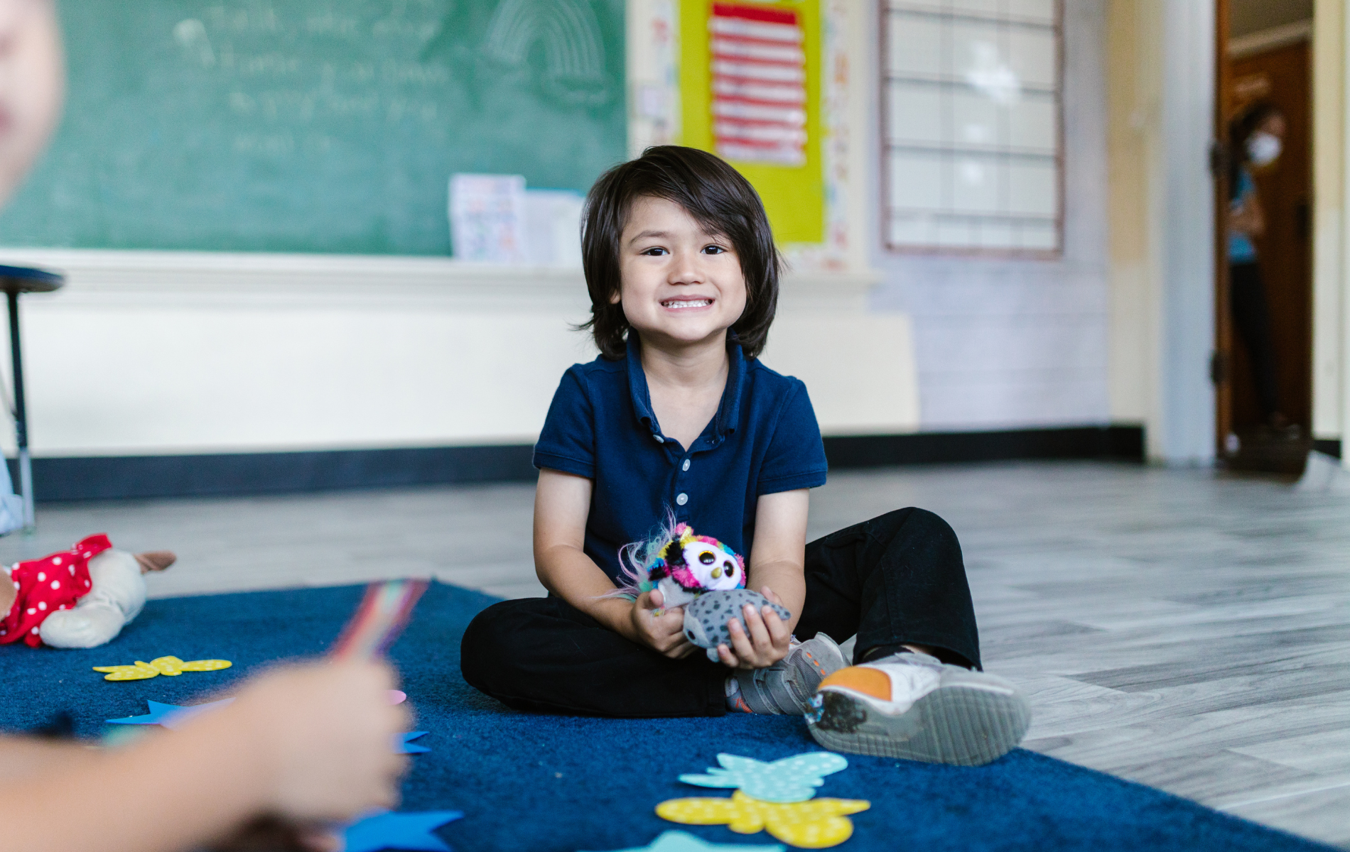 A young boy is sitting on the floor in a classroom holding a stuffed animal.