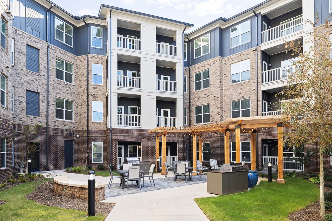 Outdoor communal courtyard with seating, a wooden pergola, and a grill in a brick apartment complex.