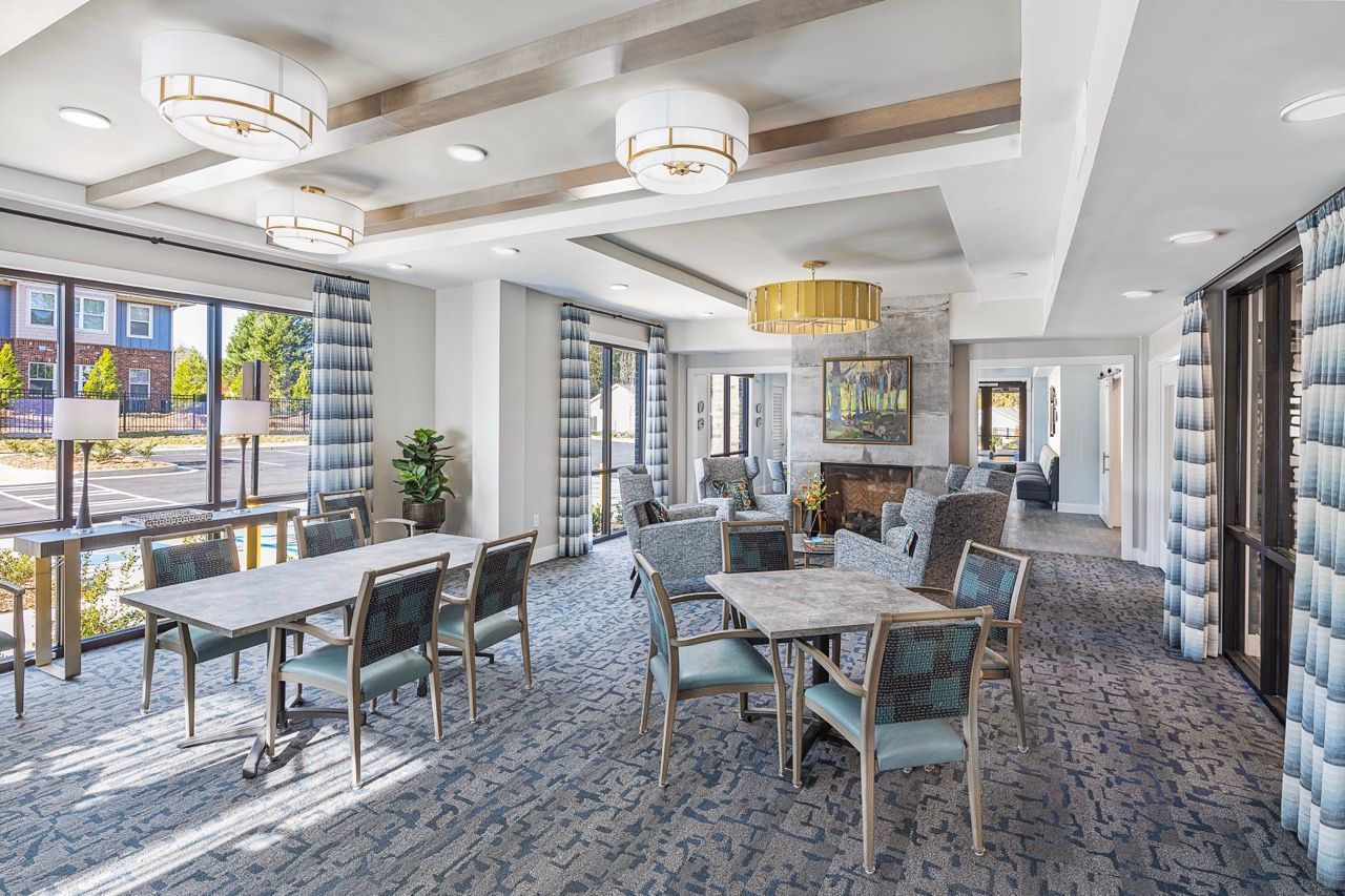 Bright resident lounge with tables, chairs, large windows, and a fireplace.