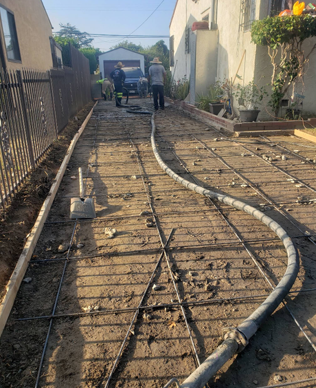 Workers laying concrete in a narrow driveway between houses, with rebar and hoses visible.