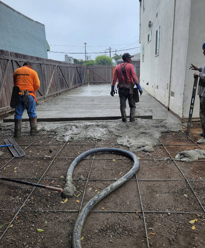 Two workers pour concrete on a wet outdoor patio beside a building, with tools and rebar in the foreground.
