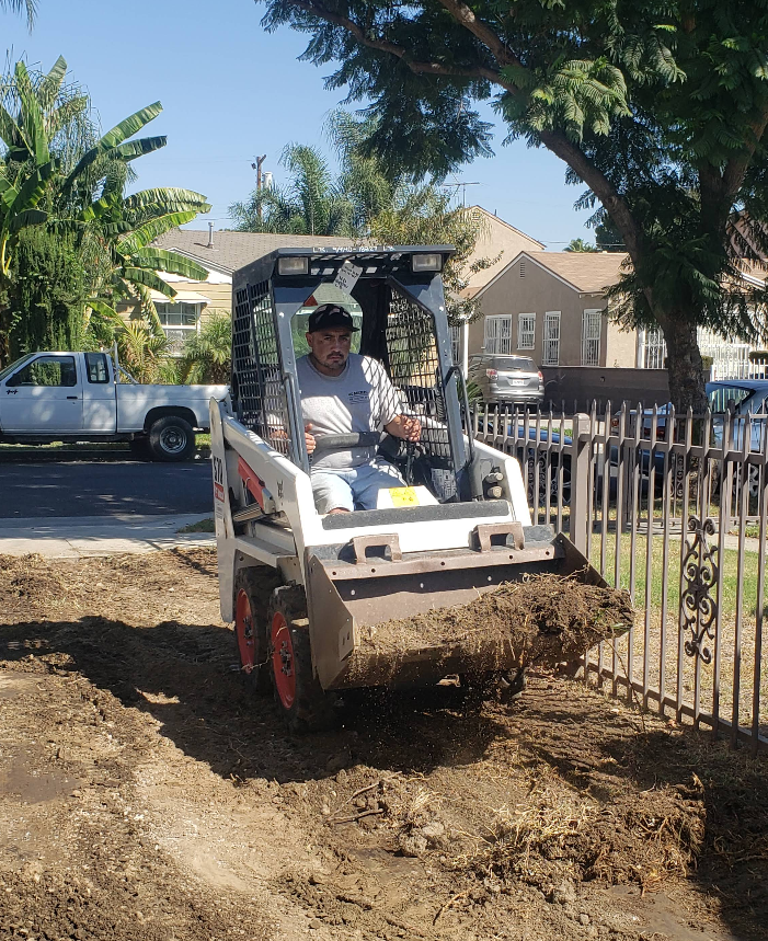 Worker driving a skid steer, scooping dirt beside a fence in a sunny residential yard