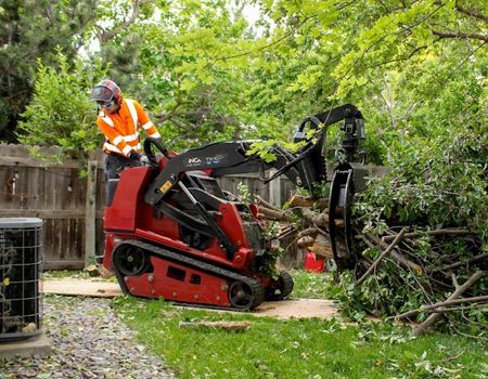 Person in orange safety gear operates a red mini skid steer, trimming branches in a backyard.