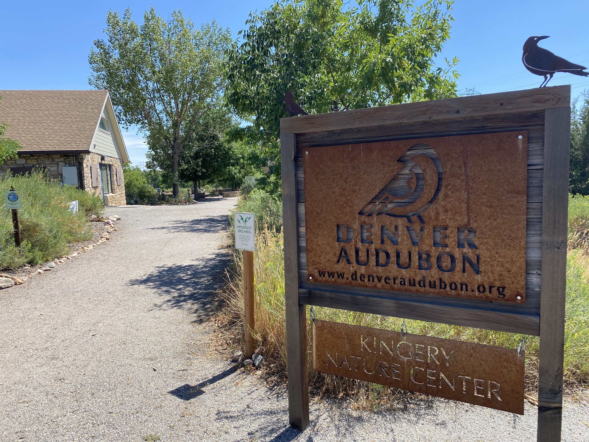 A large rust-colored metal sign reading “Denver Audubon – www.denveraudubon.org  – Kingery Nature Center” stands at the entrance of a gravel path surrounded by native plants and trees. A small bird sculpture sits on top of the wooden frame holding the sign. In the background, a stone building with a tan roof is partially visible under a clear blue sky, creating a welcoming view of the nature center.
