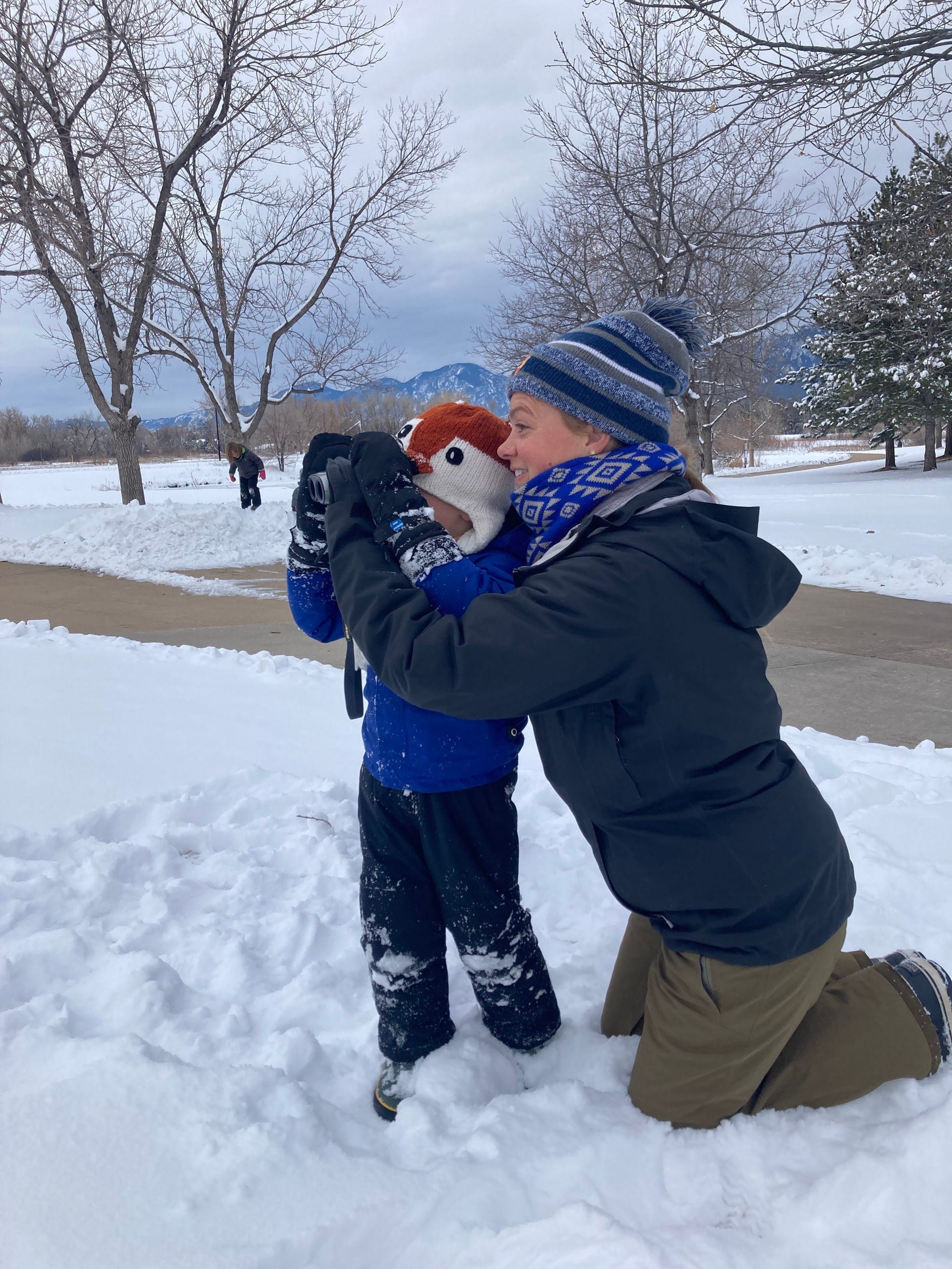Woman kneeling in snow, hugging child using binoculars. Snowy park, overcast sky.