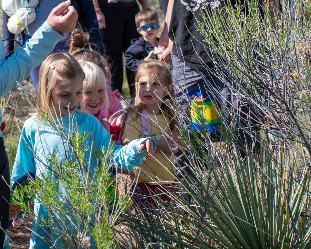Children exploring plants outdoors; a girl points with others watching.