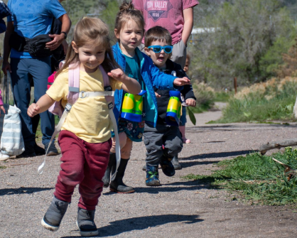 Four young children running on a dirt path, holding binoculars, sunny day.