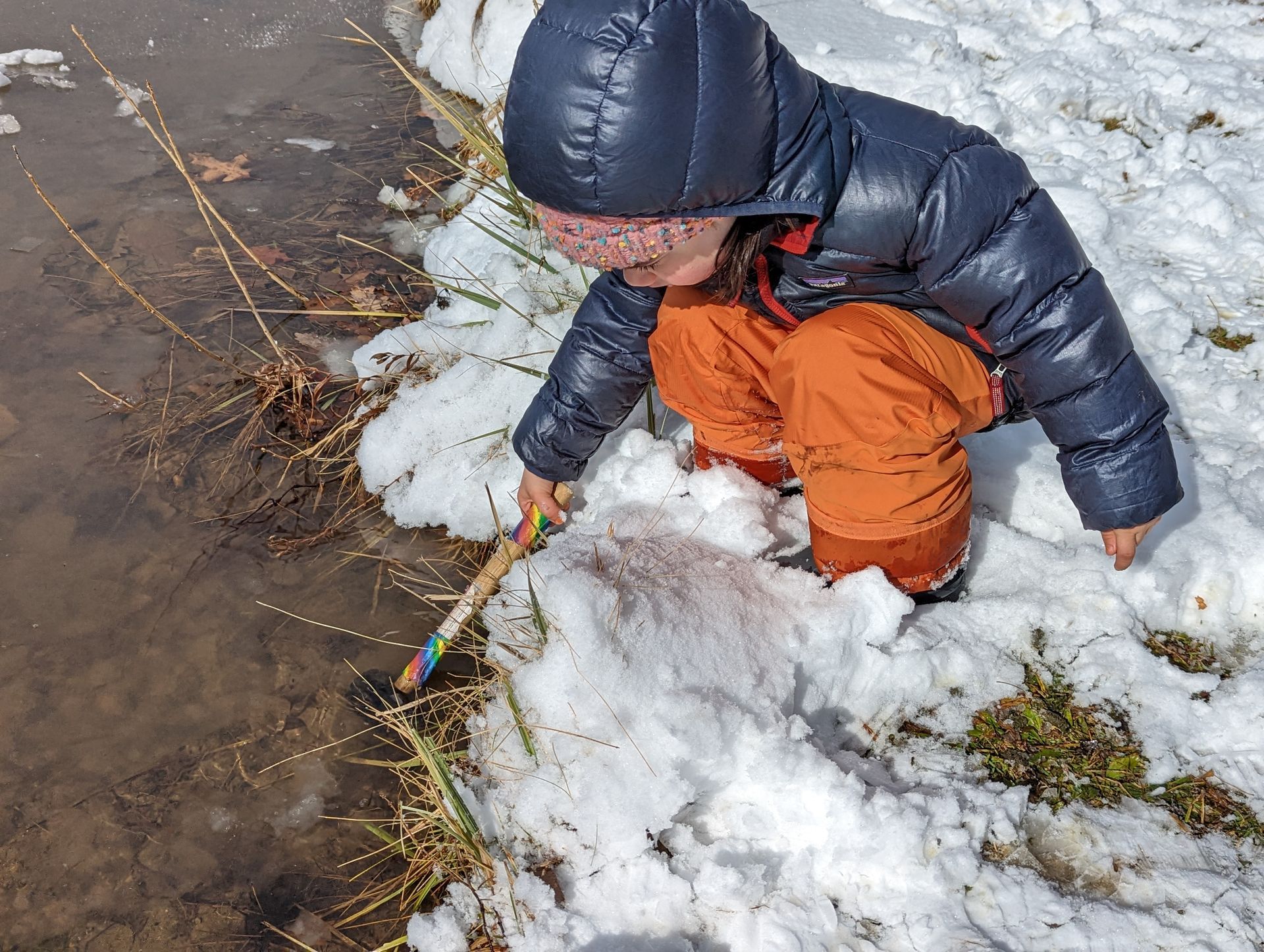 A young child dressed in a dark blue winter coat, orange snow pants, and winter boots crouches beside a snowy pond edge. The child uses a colorful stick to explore the shallow, partially thawed water surrounded by snow and dry grass. Sunlight reflects off the snow, creating a bright and curious outdoor winter scene.