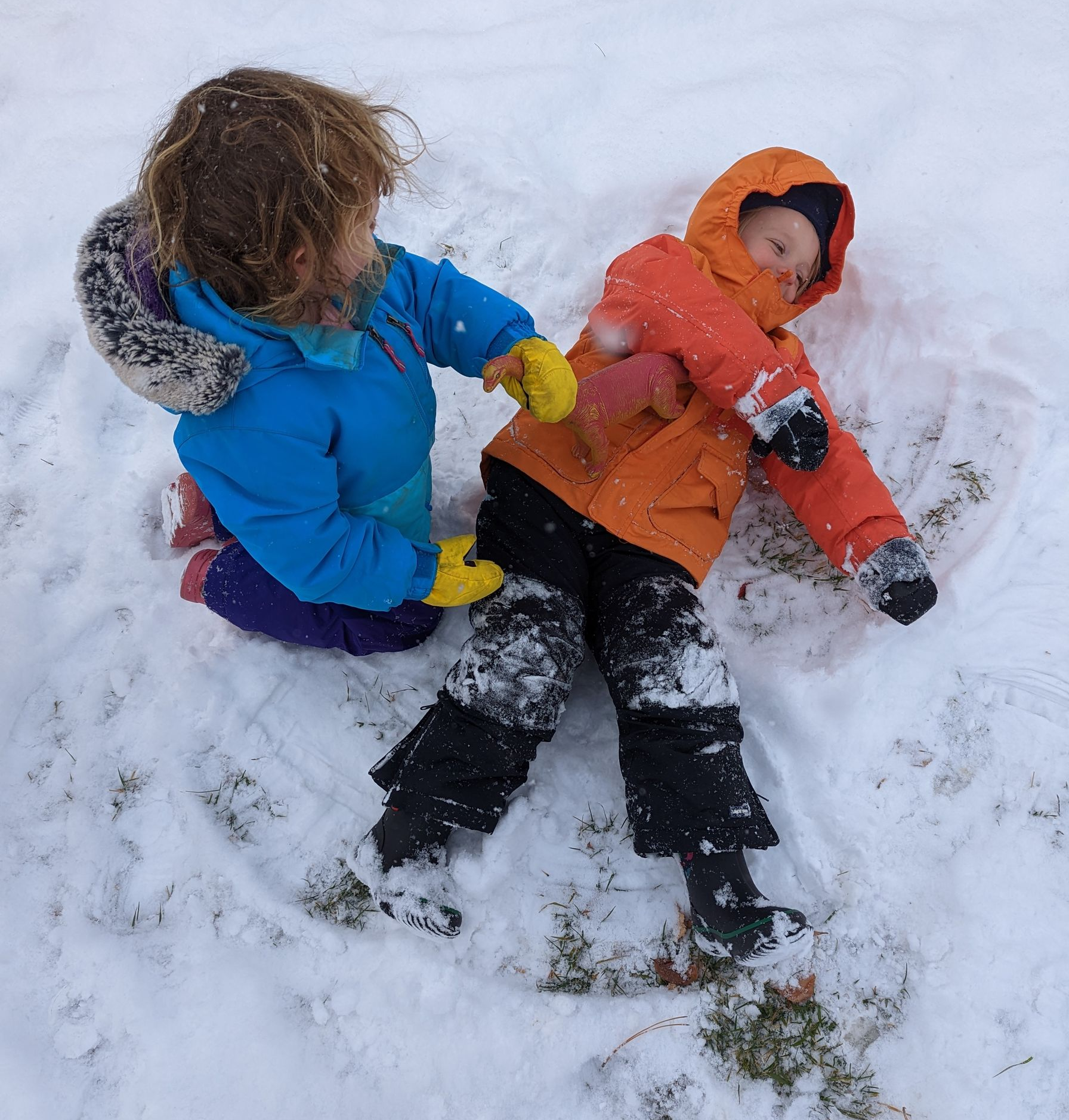 Two young children play together in the snow. One child in a blue winter coat and yellow gloves kneels beside another child lying on the ground in an orange coat and black snow pants. The child on the ground is making a snow angel while the other interacts playfully. Both are surrounded by a fresh layer of snow with small patches of grass showing through.