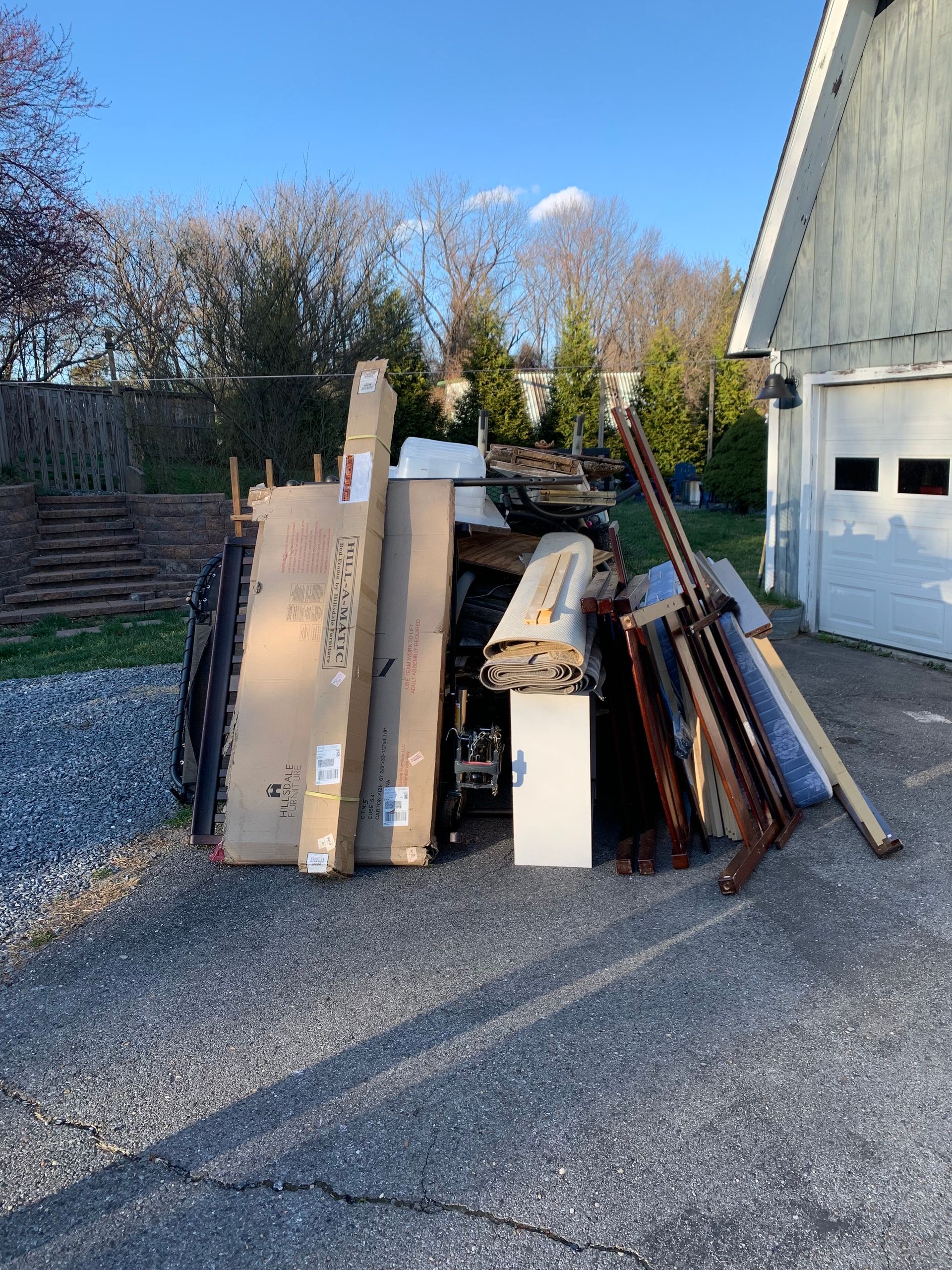 Pile of dismantled furniture and boxes on a gravel driveway next to a garage, under a sunny sky.