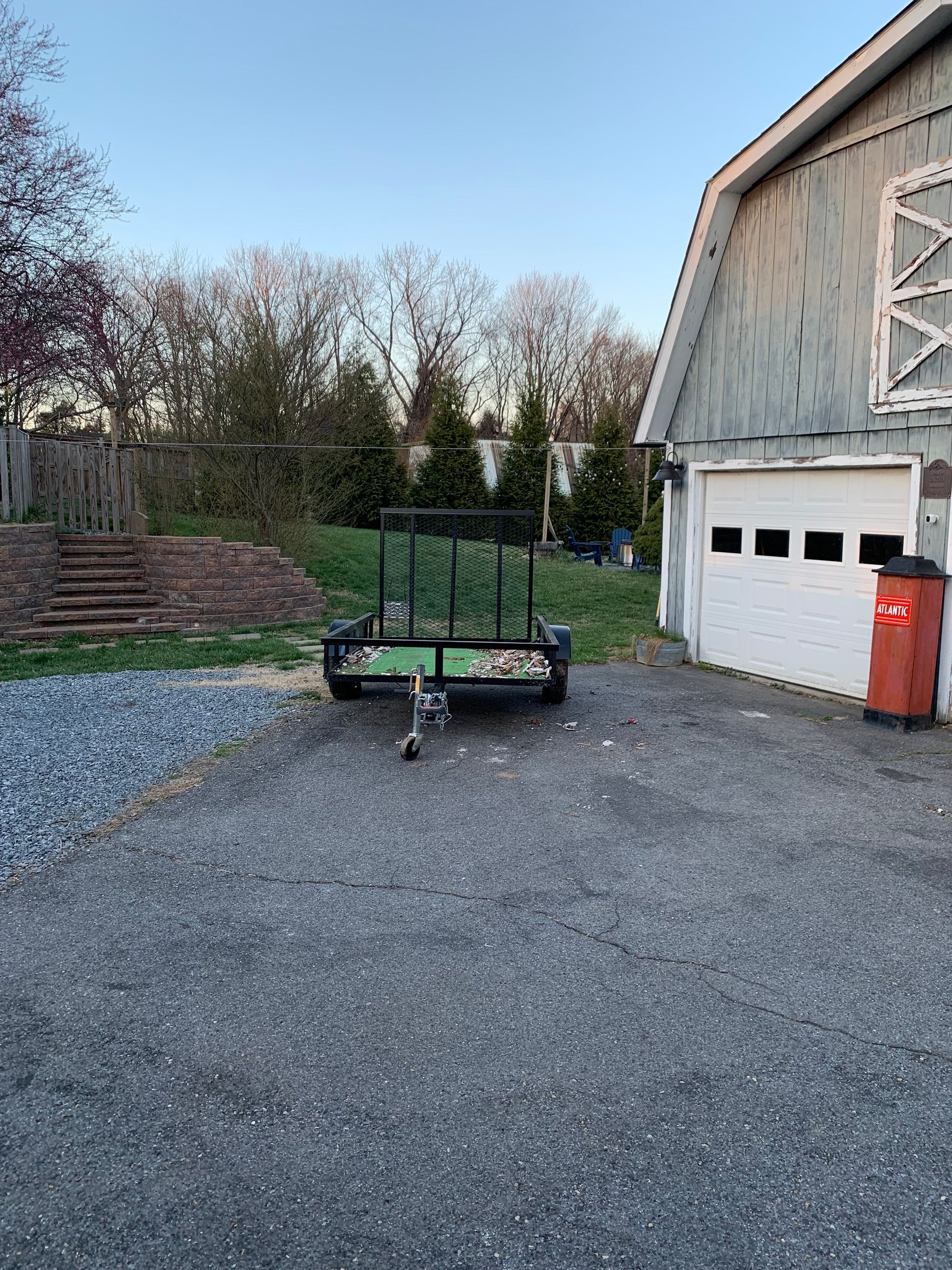 Trailer parked on gravel driveway, next to a garage and a brick staircase, with trees in the background.