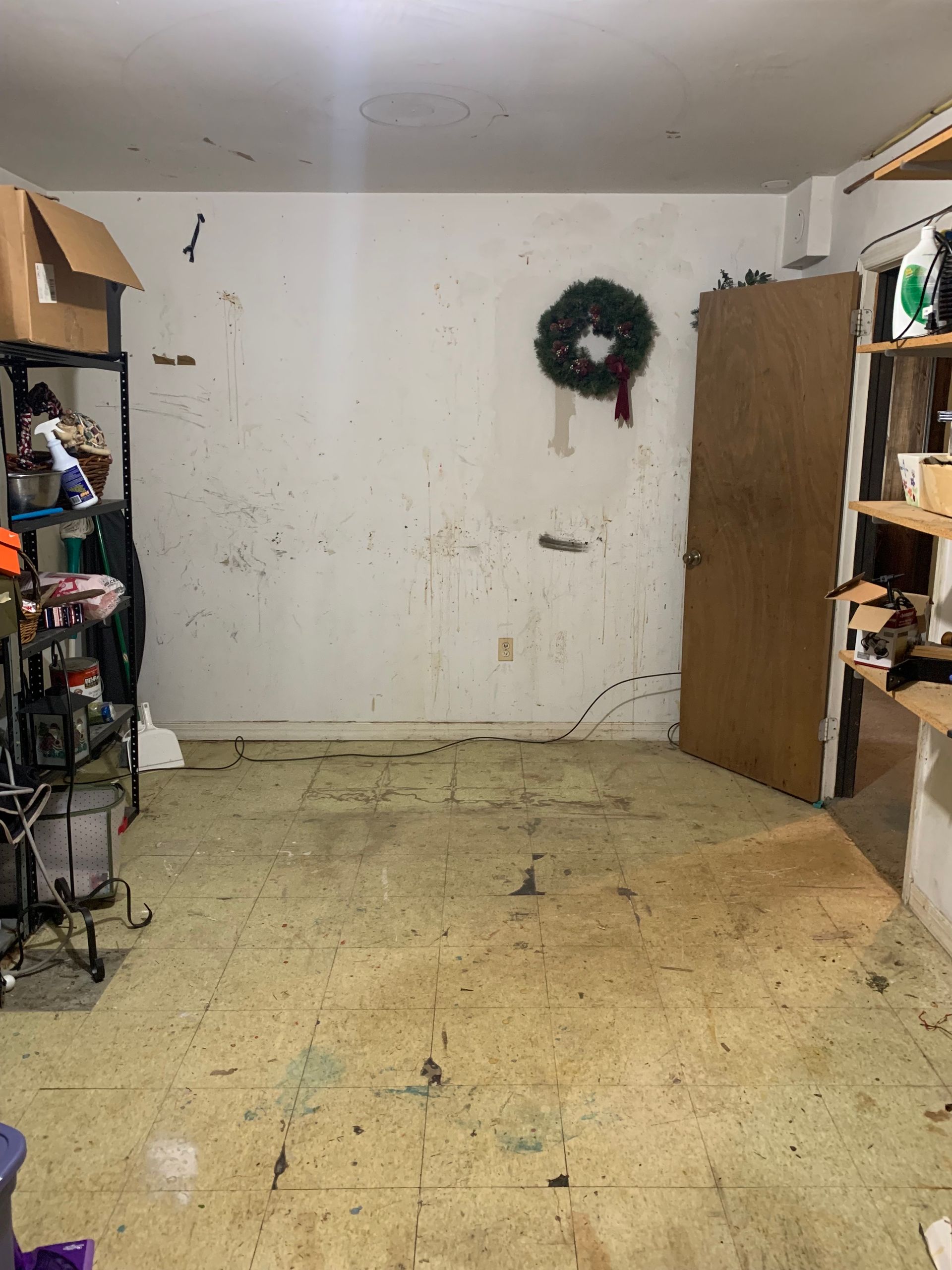 A cluttered room with dirty walls, worn floor, a door, a shelf, and a Christmas wreath.