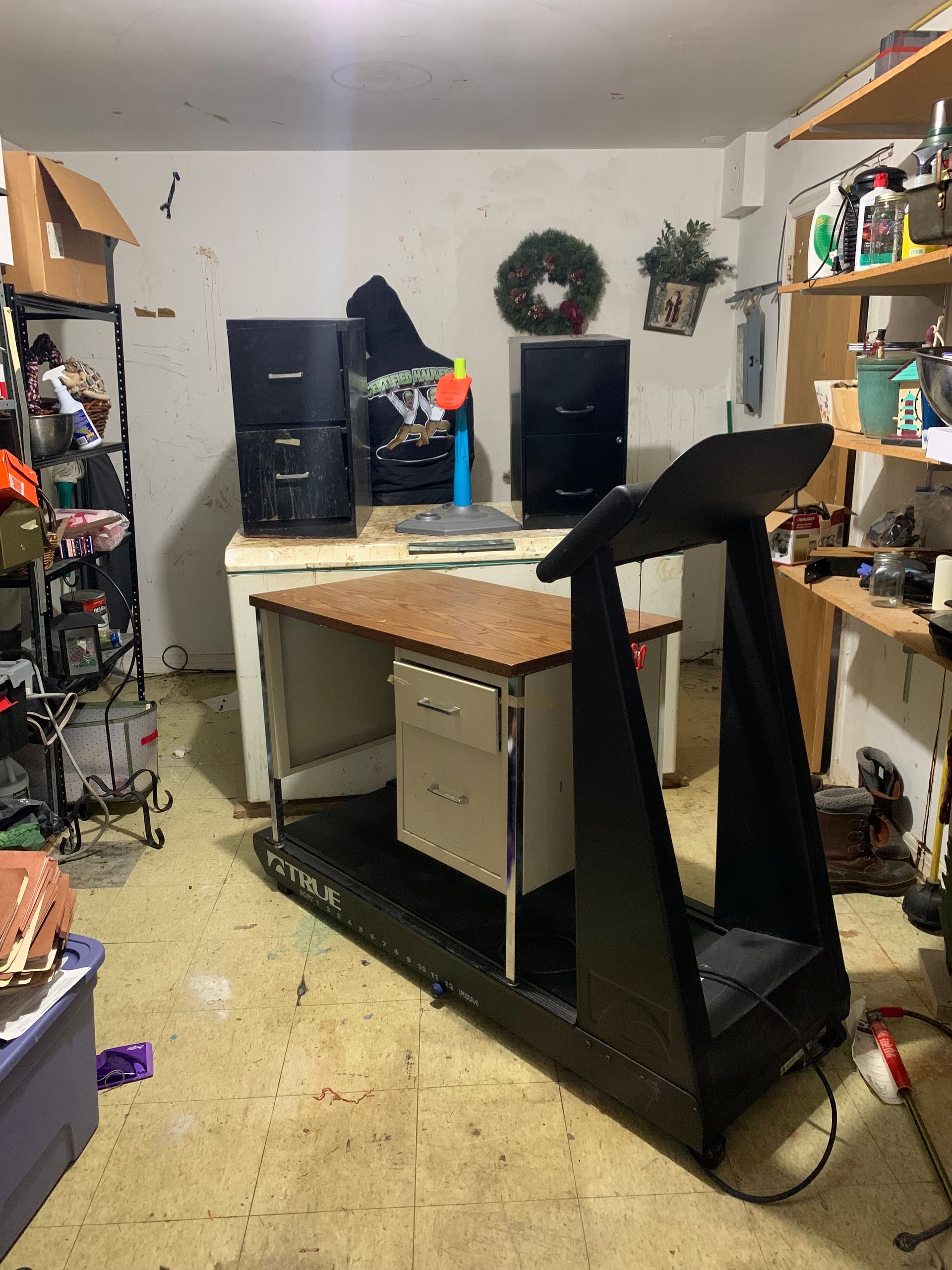 A treadmill with a desk built into it in a cluttered garage, with filing cabinets and shelves.
