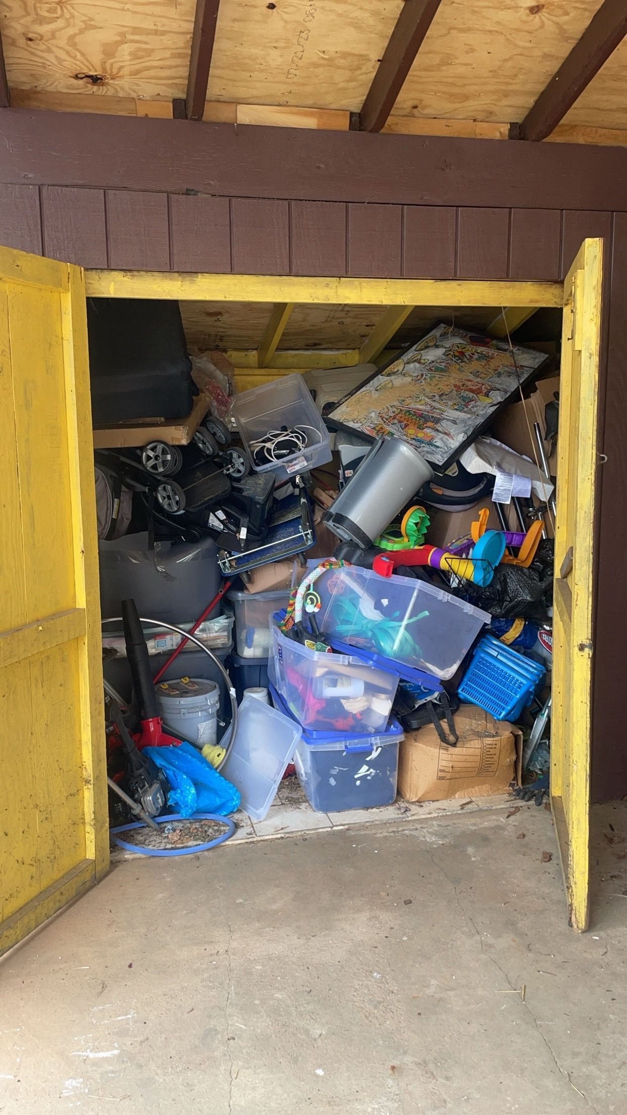 Yellow shed doors open to a cluttered interior packed with miscellaneous items, including bins, toys, and appliances.