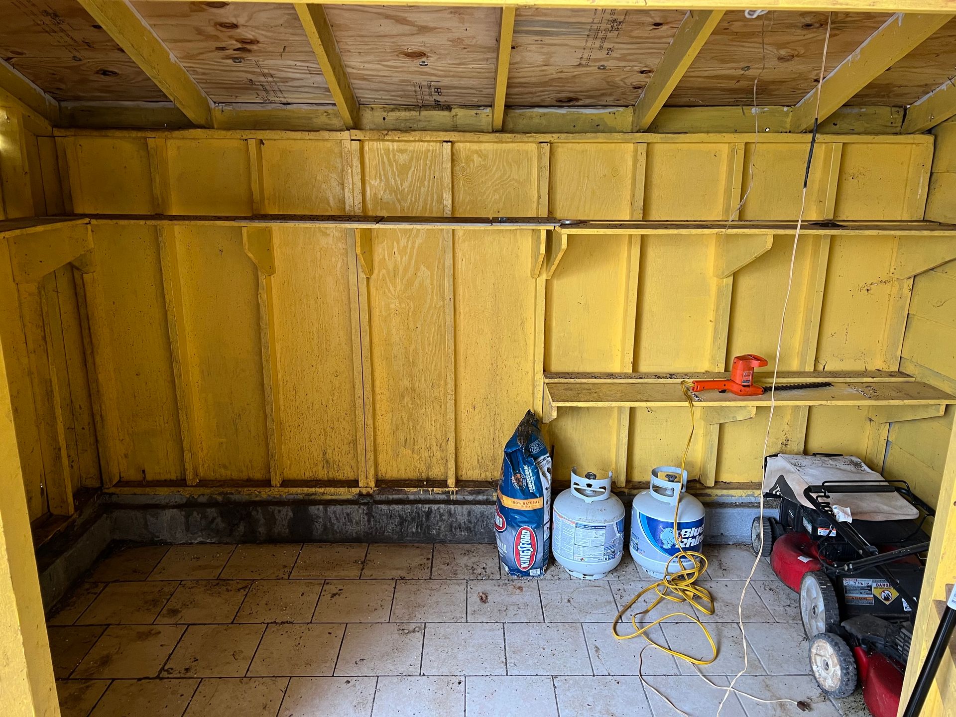Interior of a yellow shed with shelves, propane tanks, a lawnmower, and other stored items.