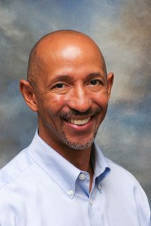 Man with shaved head smiling, wearing a light blue button-down shirt.