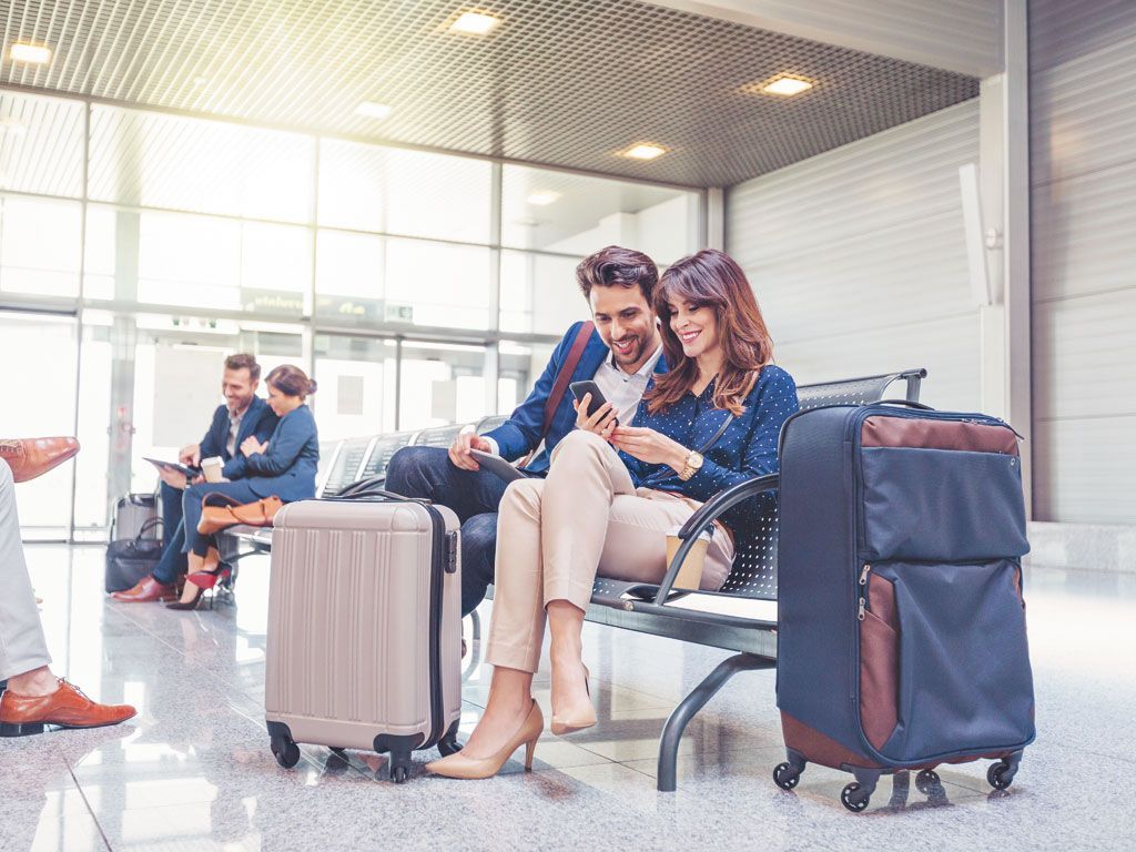 A man and a woman are sitting on a bench at an airport looking at their phones.