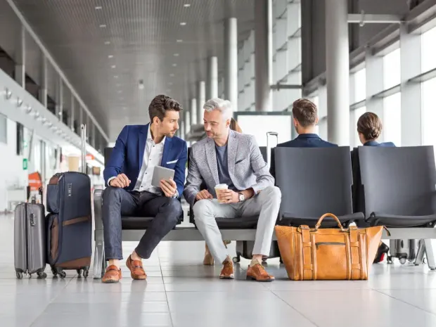 Two men are sitting on a bench at an airport looking at a tablet.