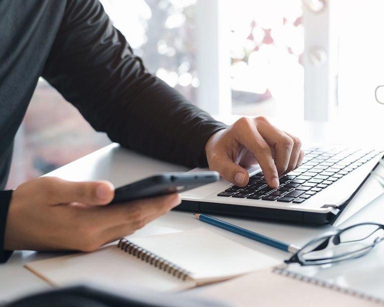 A person is typing on a laptop while holding a cell phone.