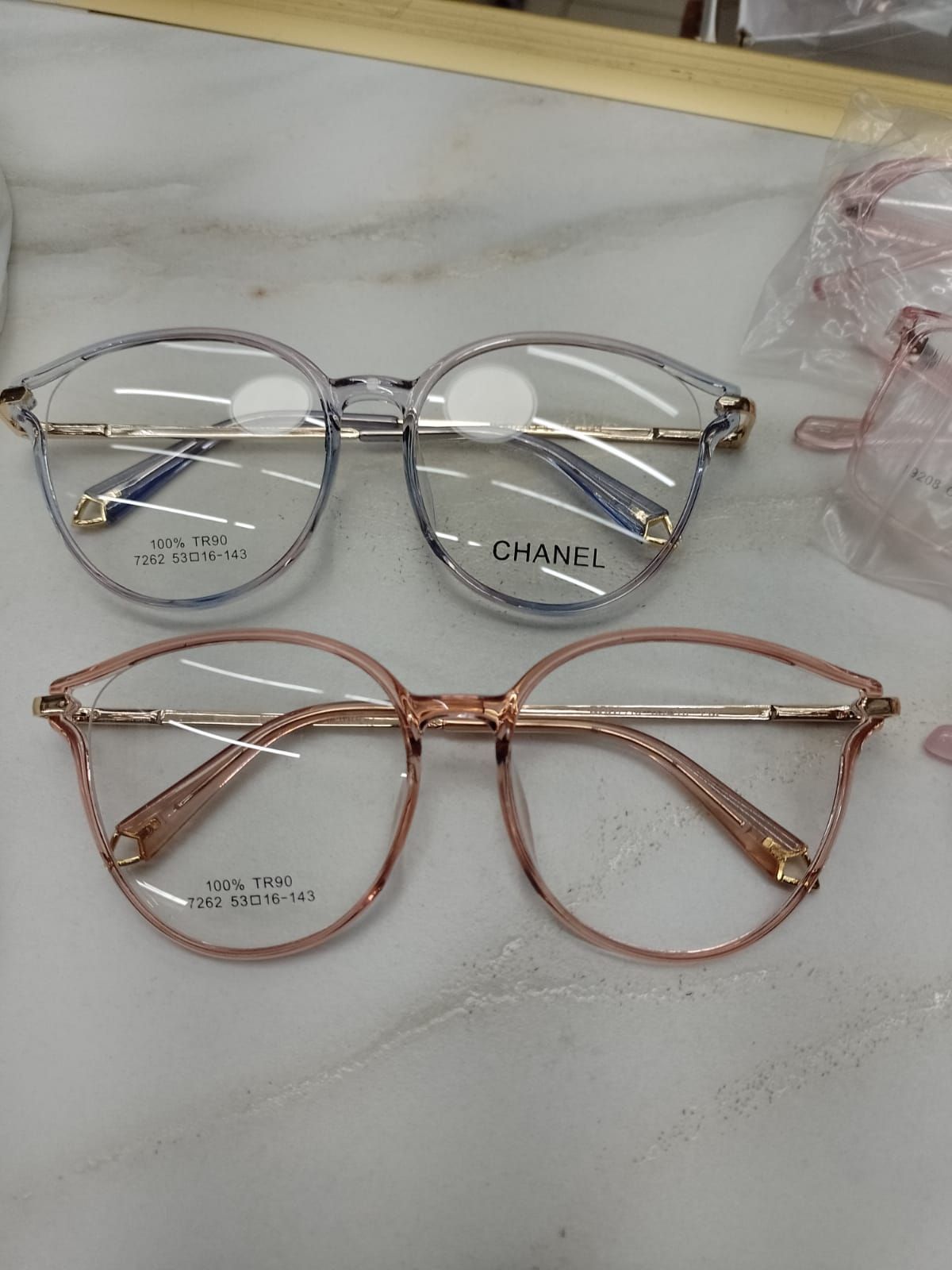 Dos pares de gafas de montura redonda, una gris translúcida y otra de oro rosa, descansan sobre una superficie blanca.