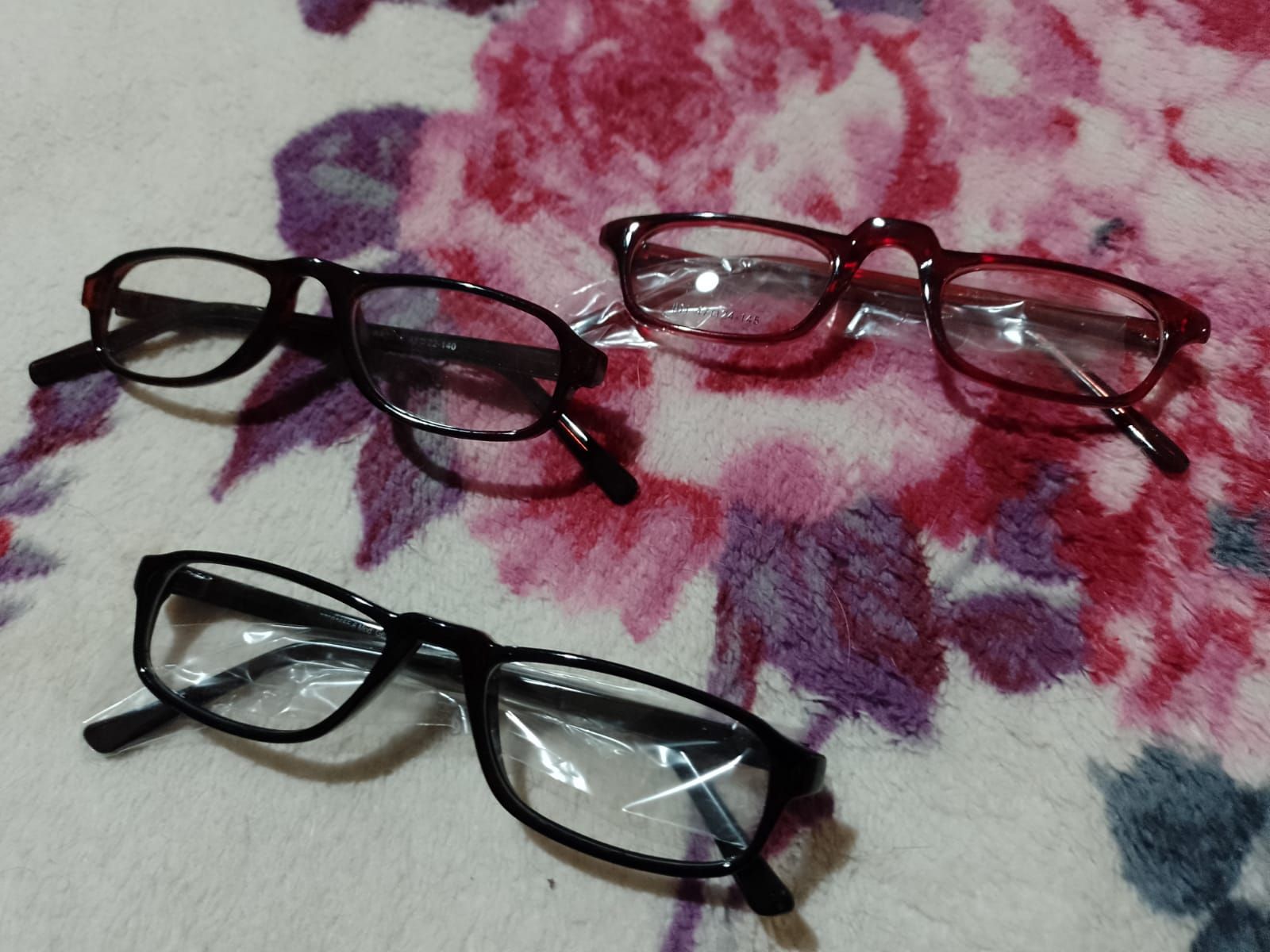 Tres pares de gafas con monturas negras y marrón rojizas descansan sobre un fondo de tela estampada con motivos florales.