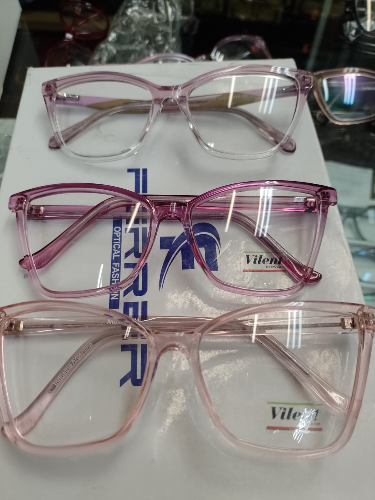 Tres pares de monturas de gafas de plástico translúcido de color claro apiladas verticalmente sobre una caja óptica de marca.