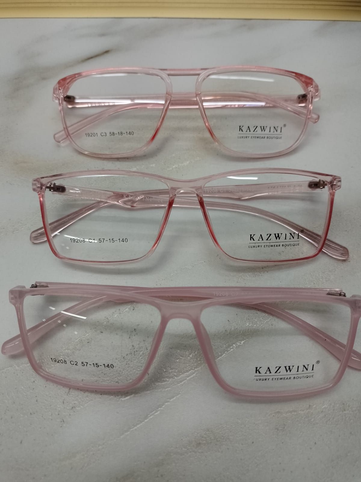 Tres pares de gafas Kazwini con montura rosa translúcida apiladas verticalmente sobre una superficie de mármol.