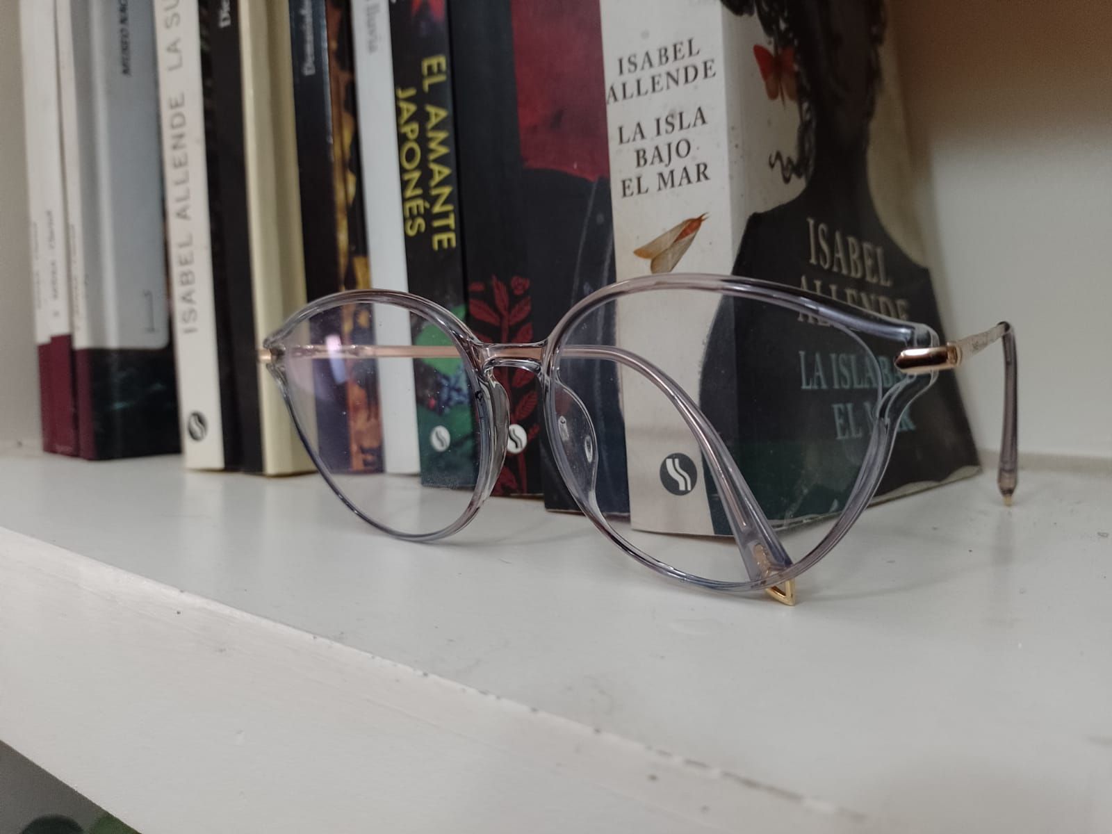 Unas gafas con montura transparente y detalles dorados descansan sobre un estante blanco delante de varios libros.