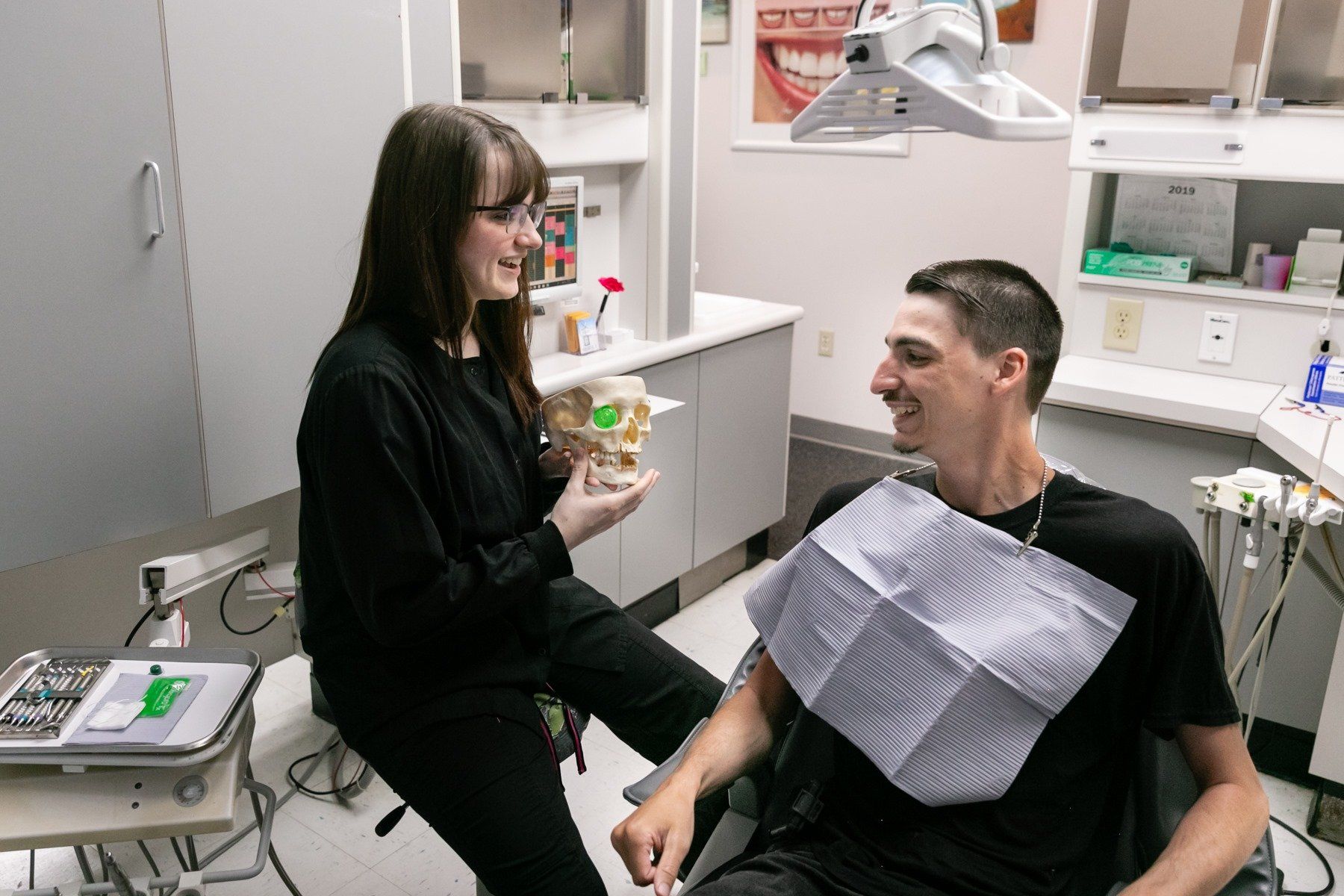 A woman is holding a model of a skull while talking to a man in a dental chair.