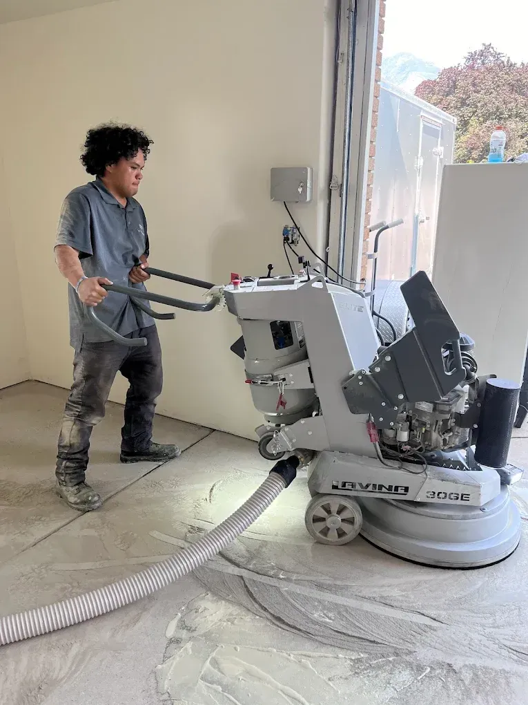 Man grinding concrete floor with large machine. White wall, open doorway, and outside foliage in background.