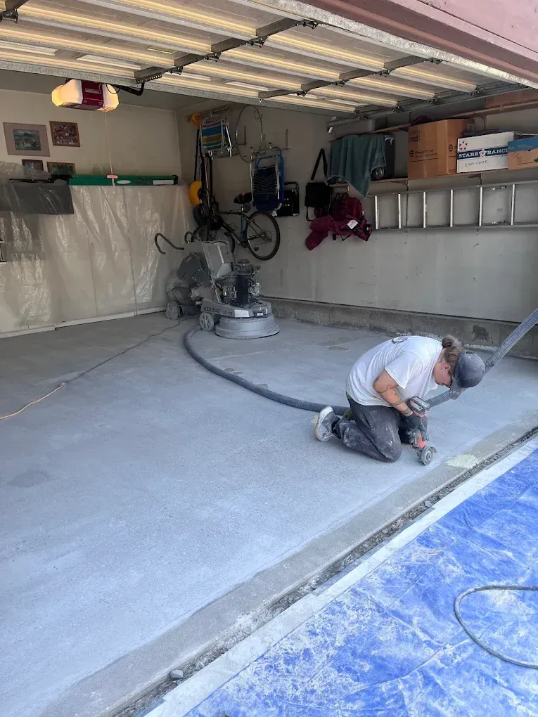 Person grinding a concrete floor in a garage; gray concrete, blue tarp, and other tools visible.