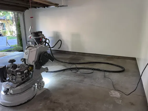 Concrete grinder in a garage, preparing floor. Gray equipment, gray floor, white wall.