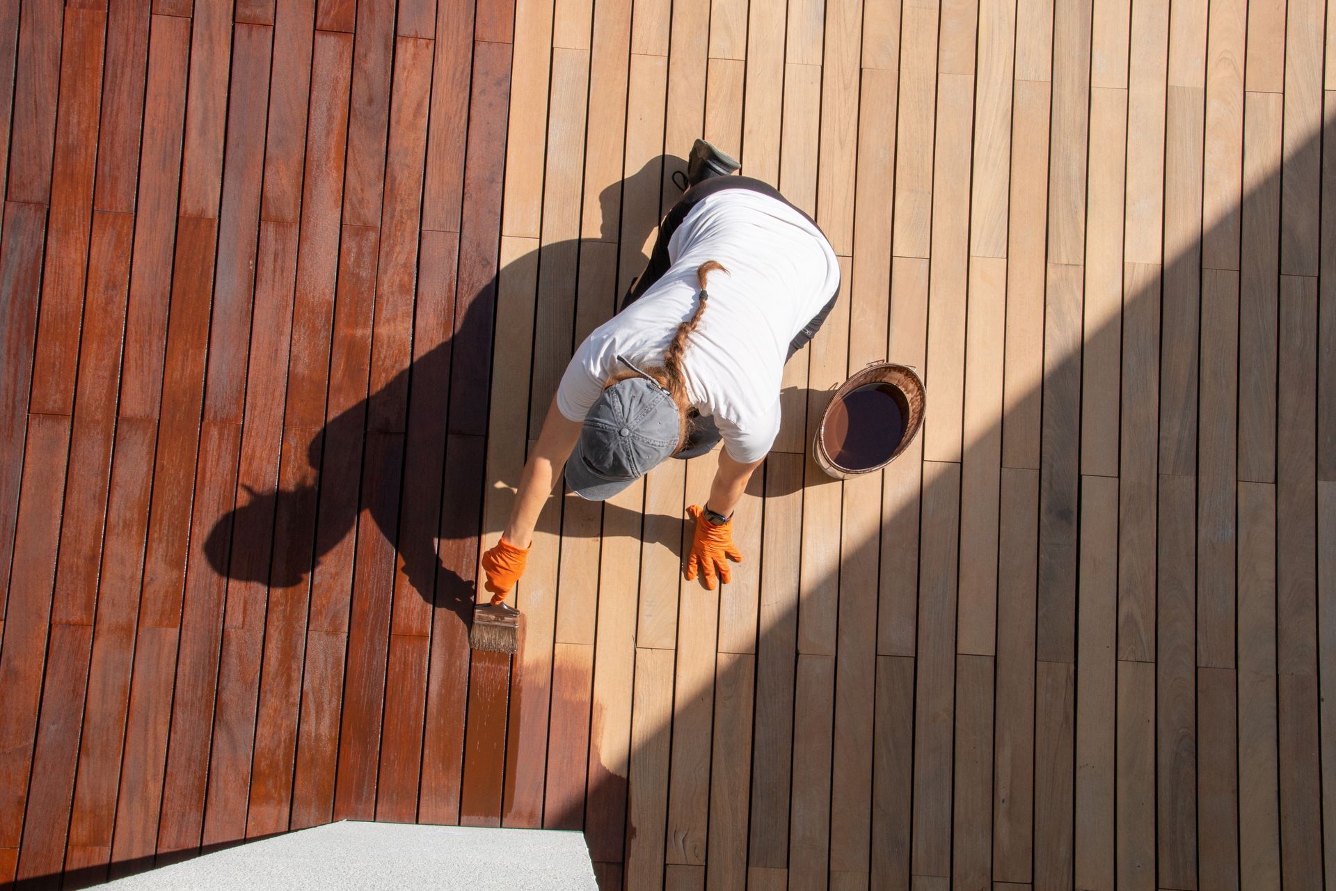Person staining a wooden deck with a reddish-brown stain, wearing gloves, and holding a brush.