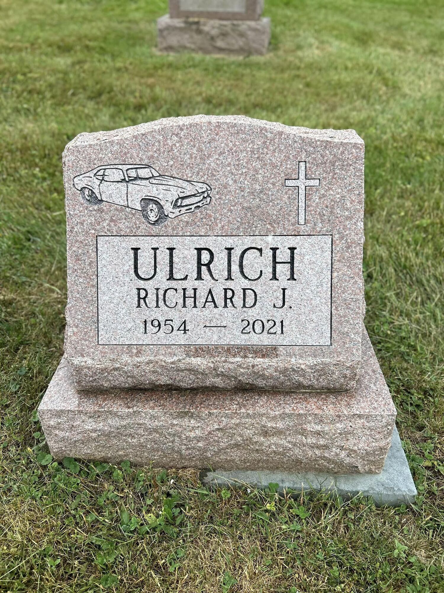 Headstone: Ulrich, Richard J. (1954-2021), with car engraving and cross, in a grassy cemetery.