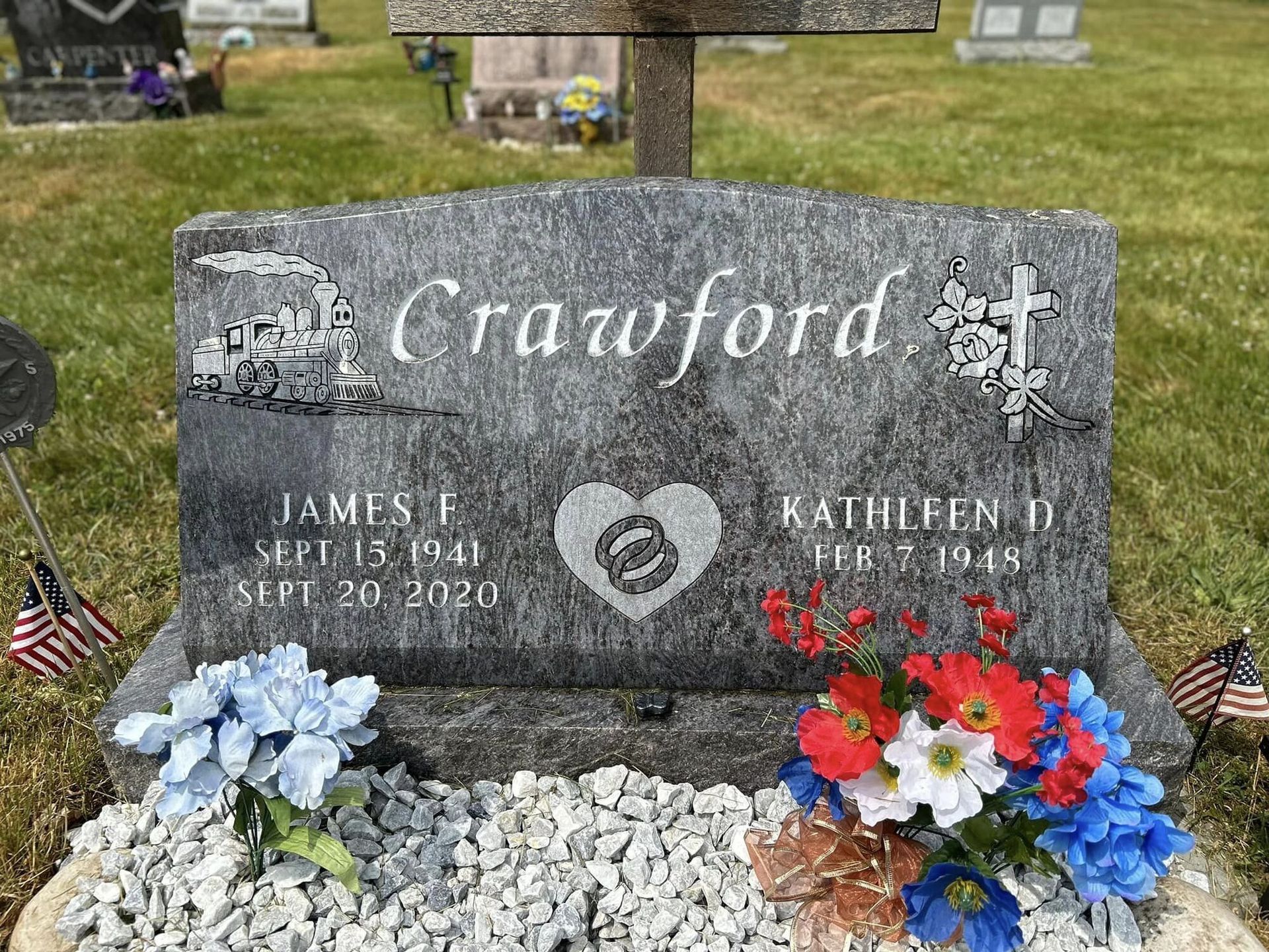 Gray headstone for Crawford family; James F. (1940-2020) & Kathleen D. (1945-). Decorated with flowers and flags.