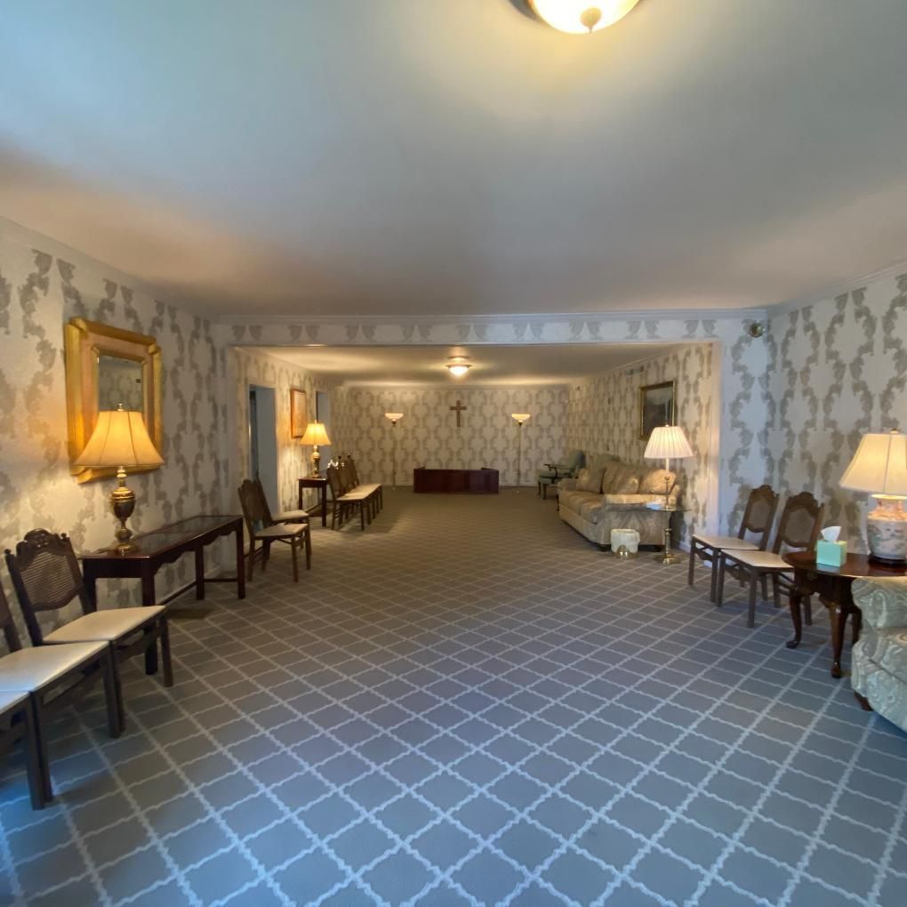 Interior of a funeral home with patterned wallpaper, chairs, lamps, and a casket.