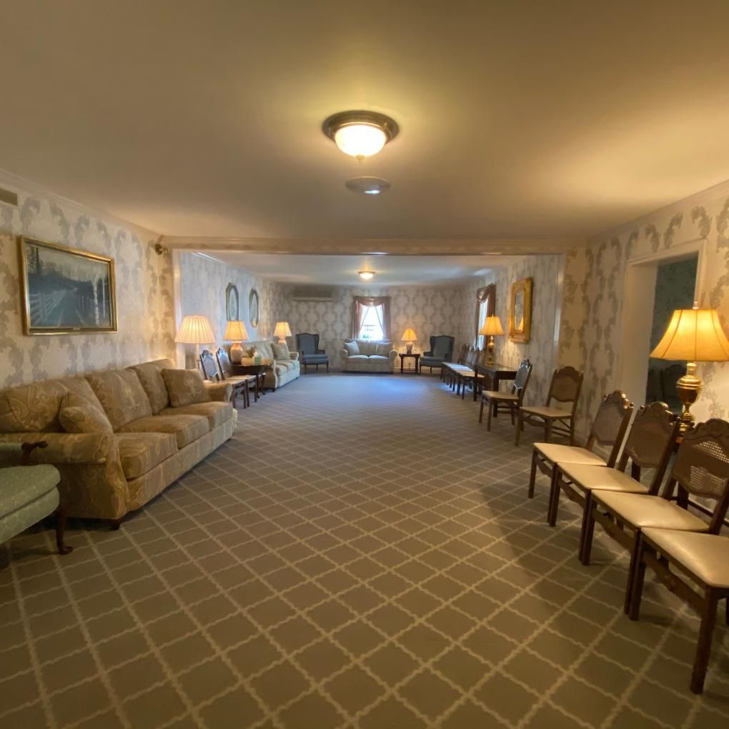 Funeral home waiting room with beige carpet, patterned wallpaper, and various seating options.