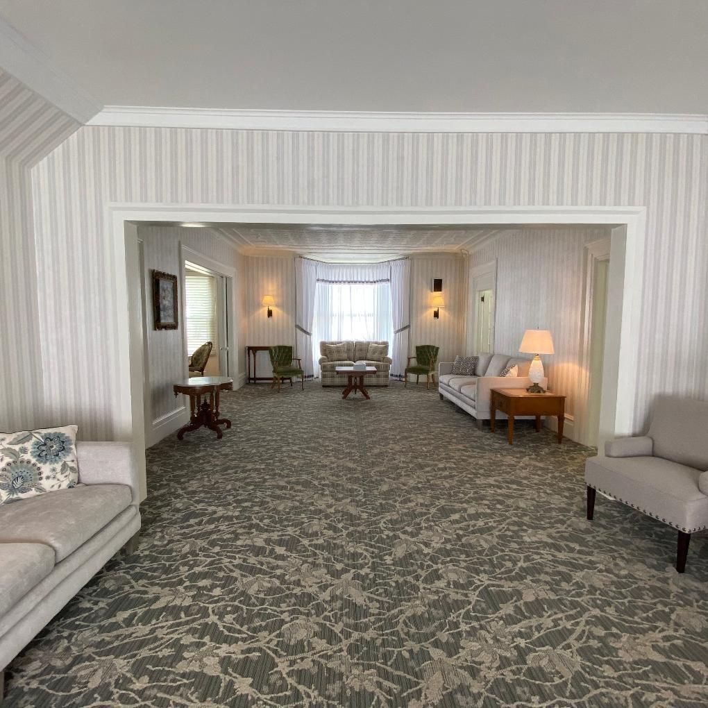 Spacious room with floral carpet, sofas, chairs, and a table; part of a well-lit, light-colored funeral home.