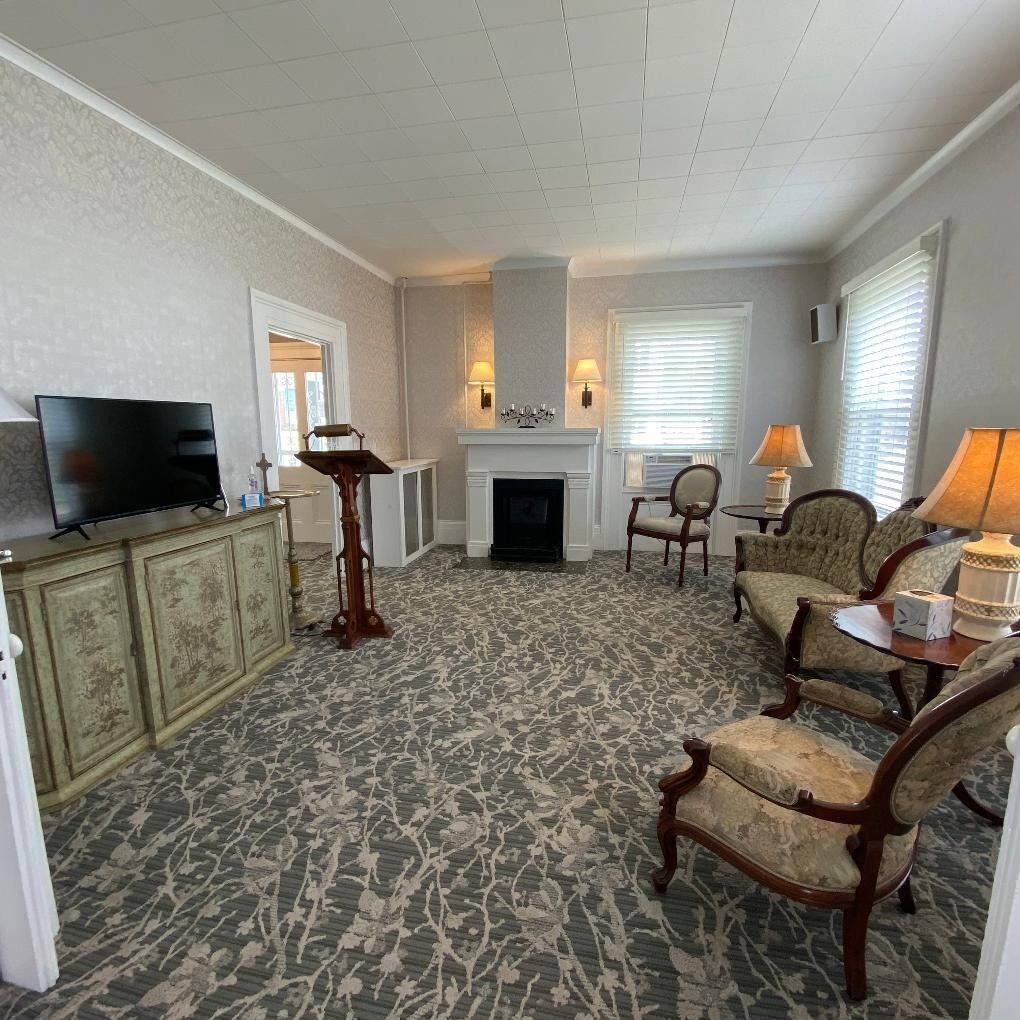 Funeral home interior; room with fireplace, chairs, and TV. Floral carpet. Light and neutral colors.