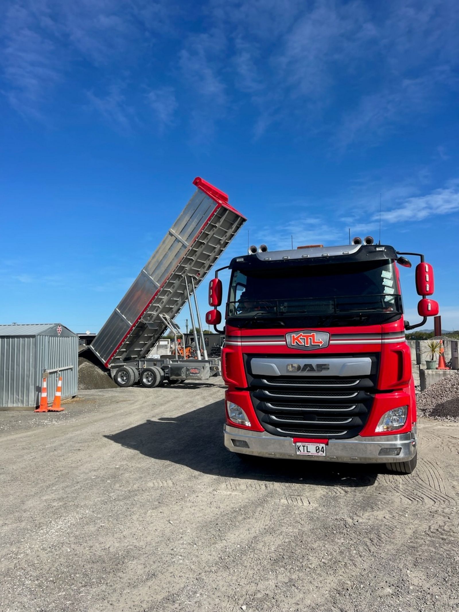 Red DAF dump truck unloading gravel at a construction site against a bright blue sky.