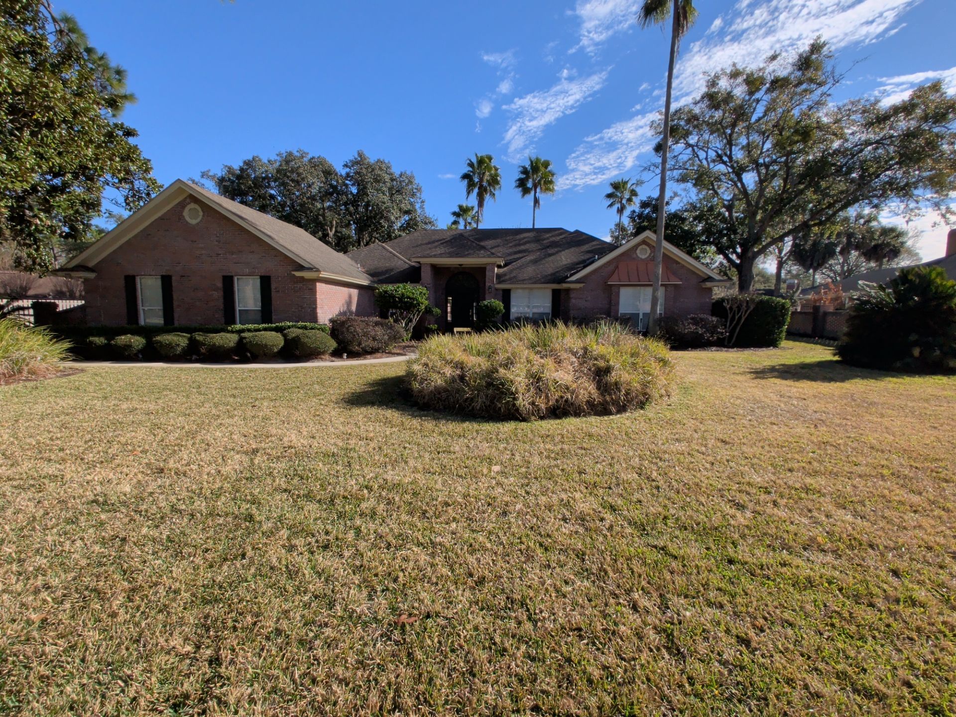 Single-story brick house with a large front lawn, bushes, and palm trees under a blue sky.