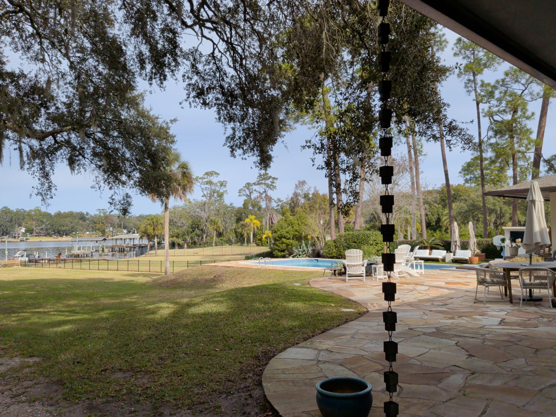 Backyard with pool, dock, and trees under a cloudy sky. Rain chain in foreground, patio furniture on stone.