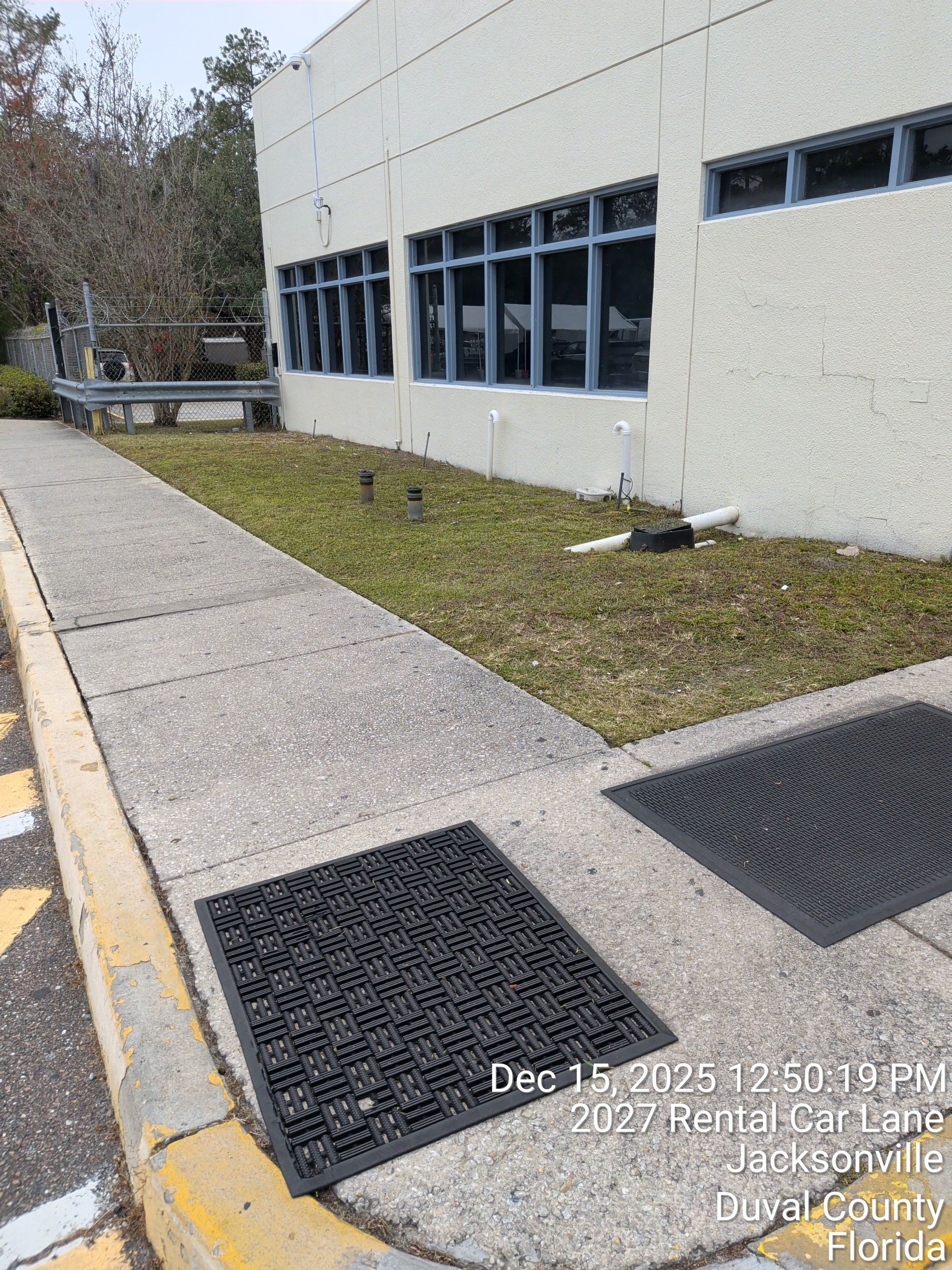 Sidewalk beside a white building with blue windows, grass strip, and black drainage mats near a curb.