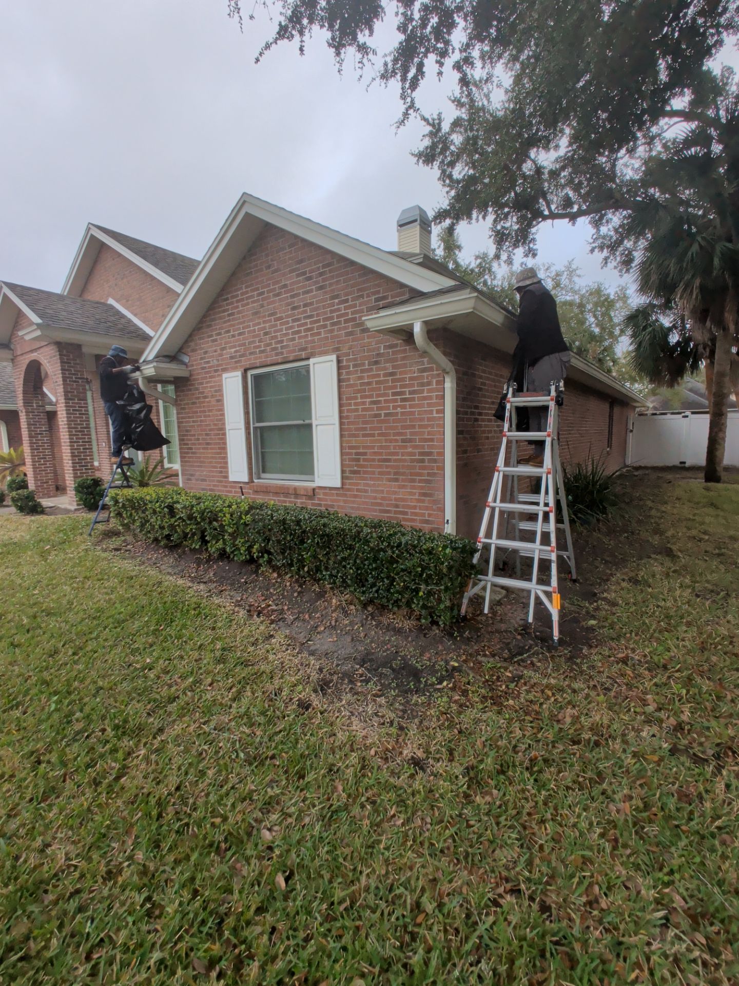 Worker on ladder repairing a brick house roof beside a green shutter, with another worker on the left